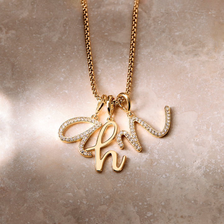 A gold initial necklace with three hanging script letters adorned with small clear stones, displayed on a textured beige surface.