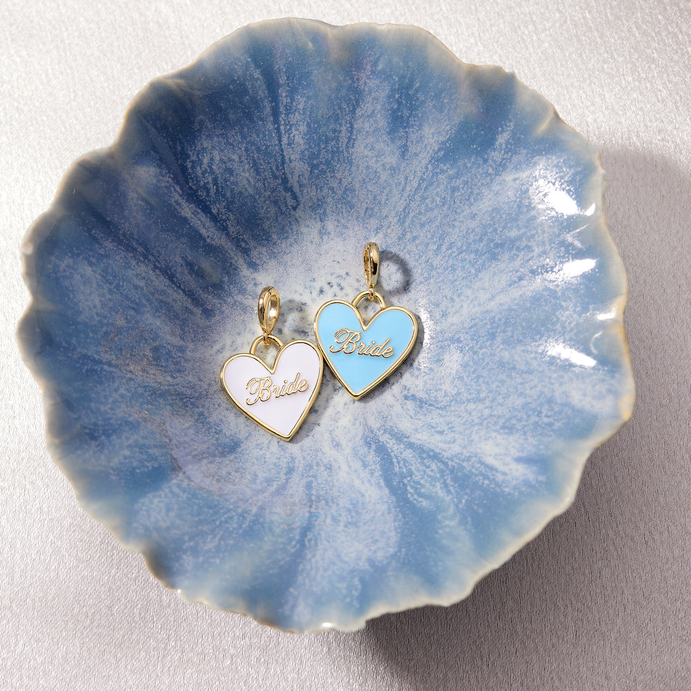  Two Effie Bride Enamel Heart charms in white and light blue enamel with script lettering, resting in a decorative ceramic bowl.