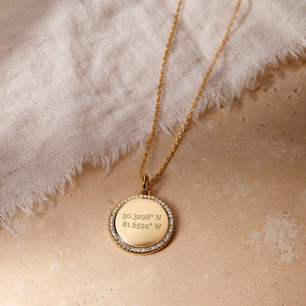  Cece Coordinate Necklace with a round pavé-bordered disc pendant engraved with custom coordinates, on a chain, laid on a stone surface.