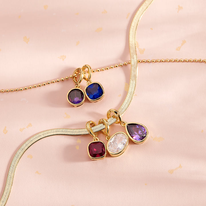 Gold-tone jewelry set featuring a delicate chain necklace with three dangling charms (square purple, round blue, oval clear) and a matching textured chain bracelet with two matching square purple and round blue charms, displayed on a soft pink patterned fabric background.