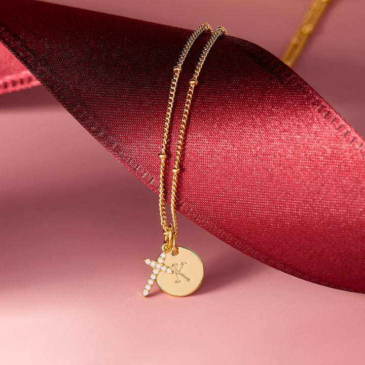 Josephine Initial Cross Pendant necklace with a pavé CZ cross charm and engraved round disc charm, draped over a red ribbon.