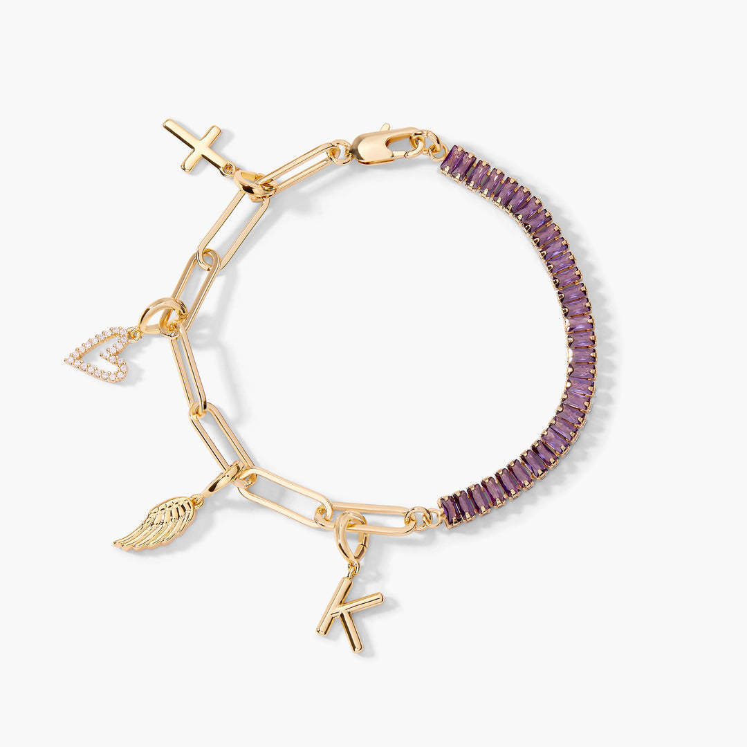 Gold-tone Priya Birthstone Charm Bracelet with purple baguette stones on one half and a chain with a cross, pavé heart, wing, and initial letter charm on the other.